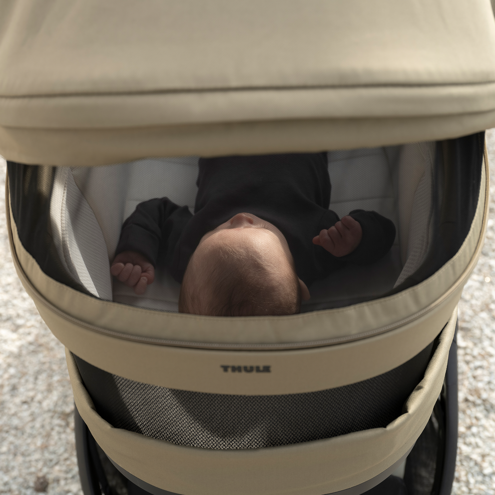 Baby lying inside a Thule Charm stroller bassinet with a mesh cover partially closed.