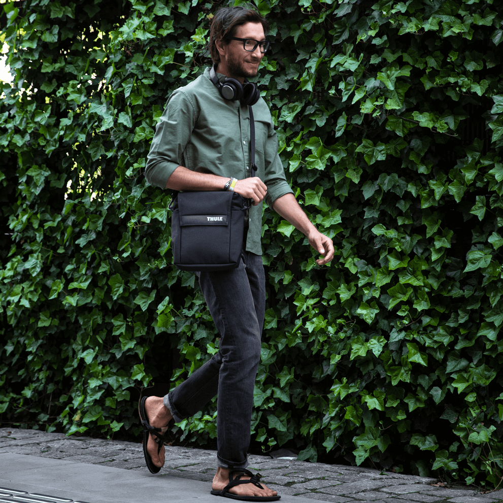 A man walks down the street with headphones, a green shirt and a Thule Paramount crossbody tote.