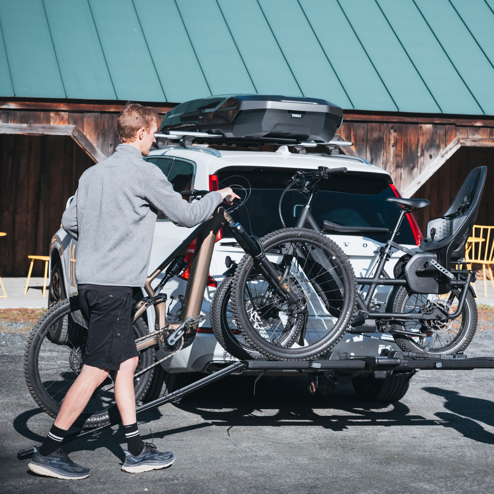 A man is using the Thule foldable loading ramp to load a bike on the Thule Vero hitch bike rack mounted on the back of a car, carrying another bike, parked in an outdoor setting.