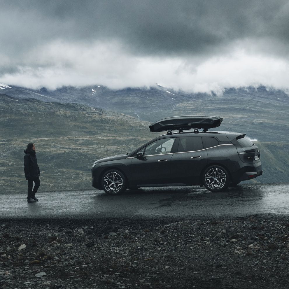 A person walks towards a vehicle parked next to the mountains with a black Thule Motion 3 roof box.
