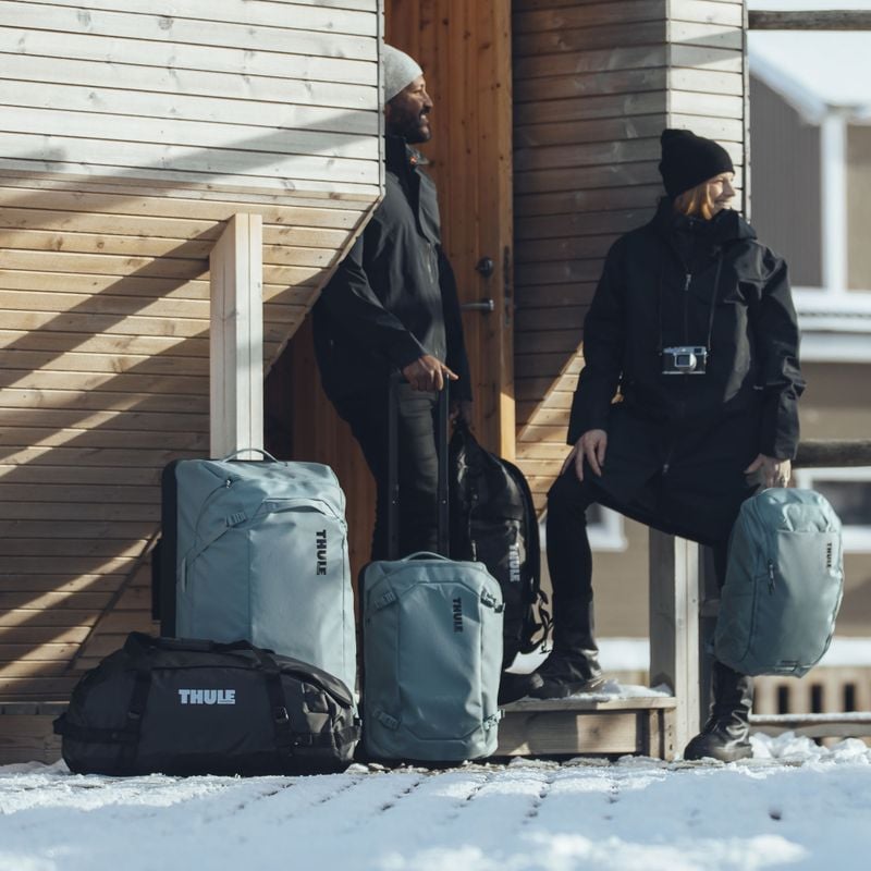 Two people stand next to a wooden structure with blue and black Thule Chasm duffels, suitcases and backpacks.