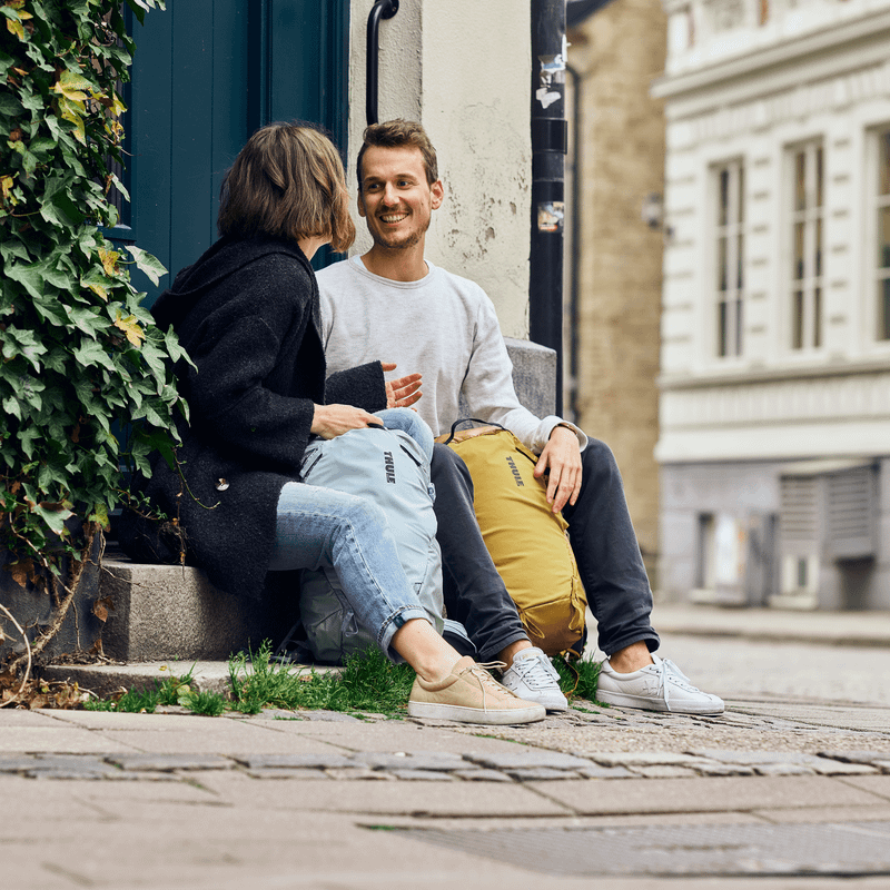A man and woman sit on a ledge at the corner of the street chatting with Thule Stir 18L backpacks.