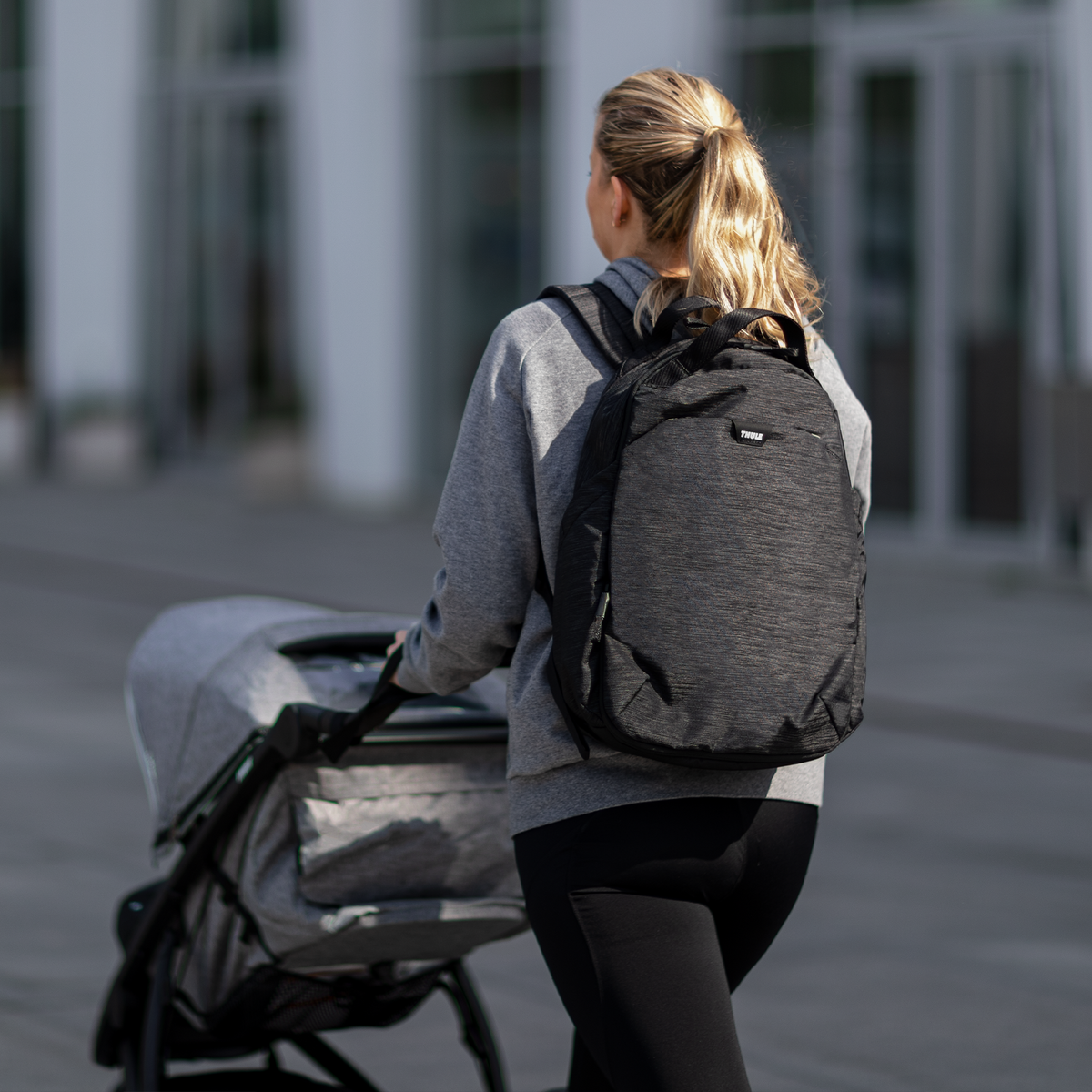 A woman walks down the street with her gray stroller and a black Thule Changing Backpack.