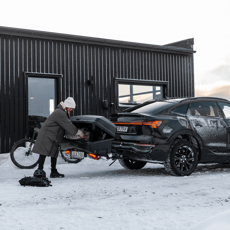 A person is putting cargo in a Thule Santu cargo box onto a Thule Epos bike carrier mounted on a car, in a snowy setting.