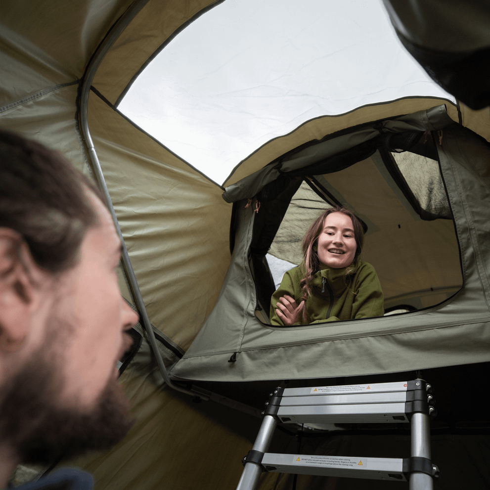 A woman looks out of a roof top tent and greets a bearded man.