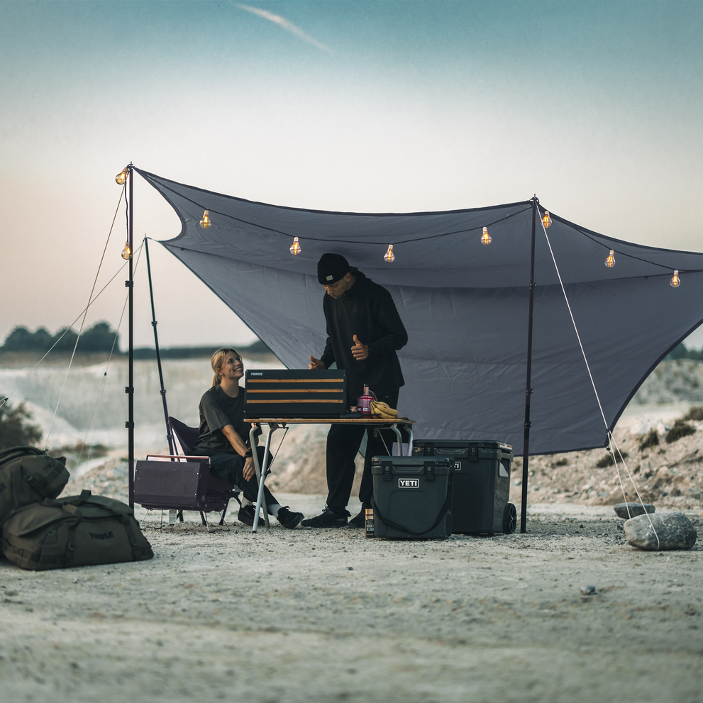 A woman and a man are enjoying an outdoors summer camping environment with shelter from the weather protective Thule Starset tarp.