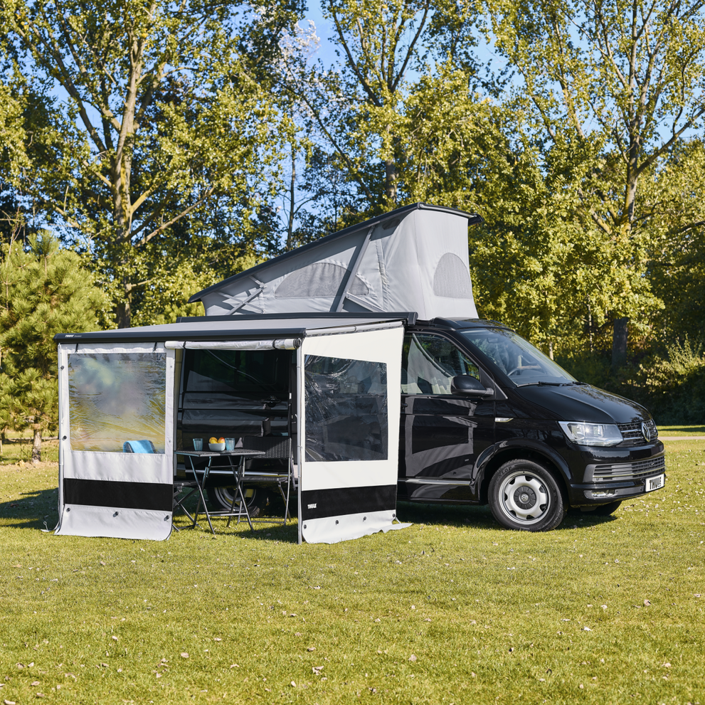 Compact van with pop-up roof and open Thule awning with panel set set up with a door opening on a grass field in the sun.