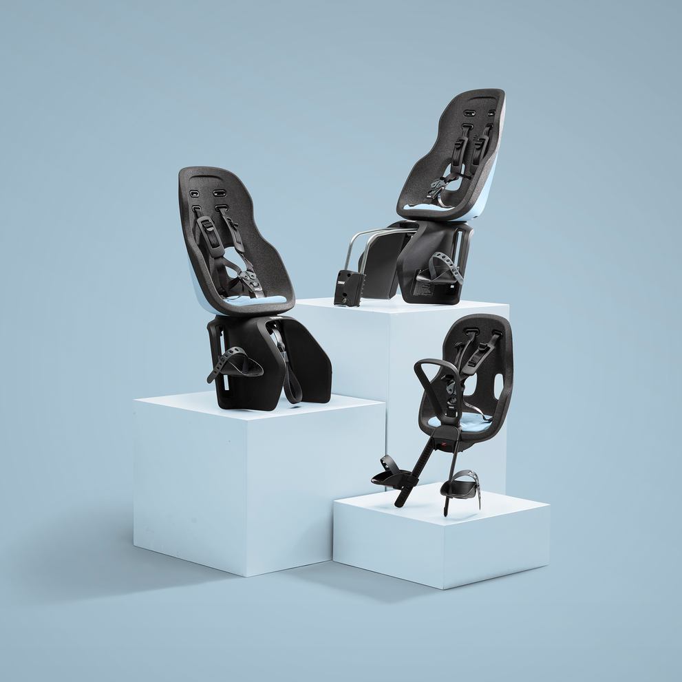 Três cadeiras de bicicleta para crianças são exibidas em diferentes alturas em pedestais brancos contra um fundo verde neutro. Cada cadeira apresenta um design elegante com um cinto de segurança ajustável e alças para os pés