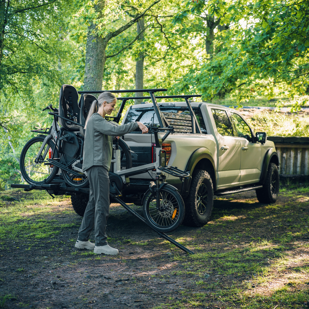 A woman is using the Thule foldable loading ramp to unload a heavy e-bike of the Thule Vero hitch bike rack mounted on the back of a truck, carrying another bike, parked in an outdoor setting.