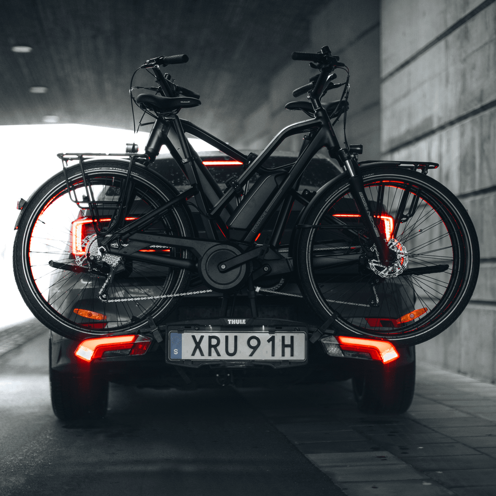 Under an overpass, a black car is parked with a black bike on a black Thule Epos hitch bike rack.