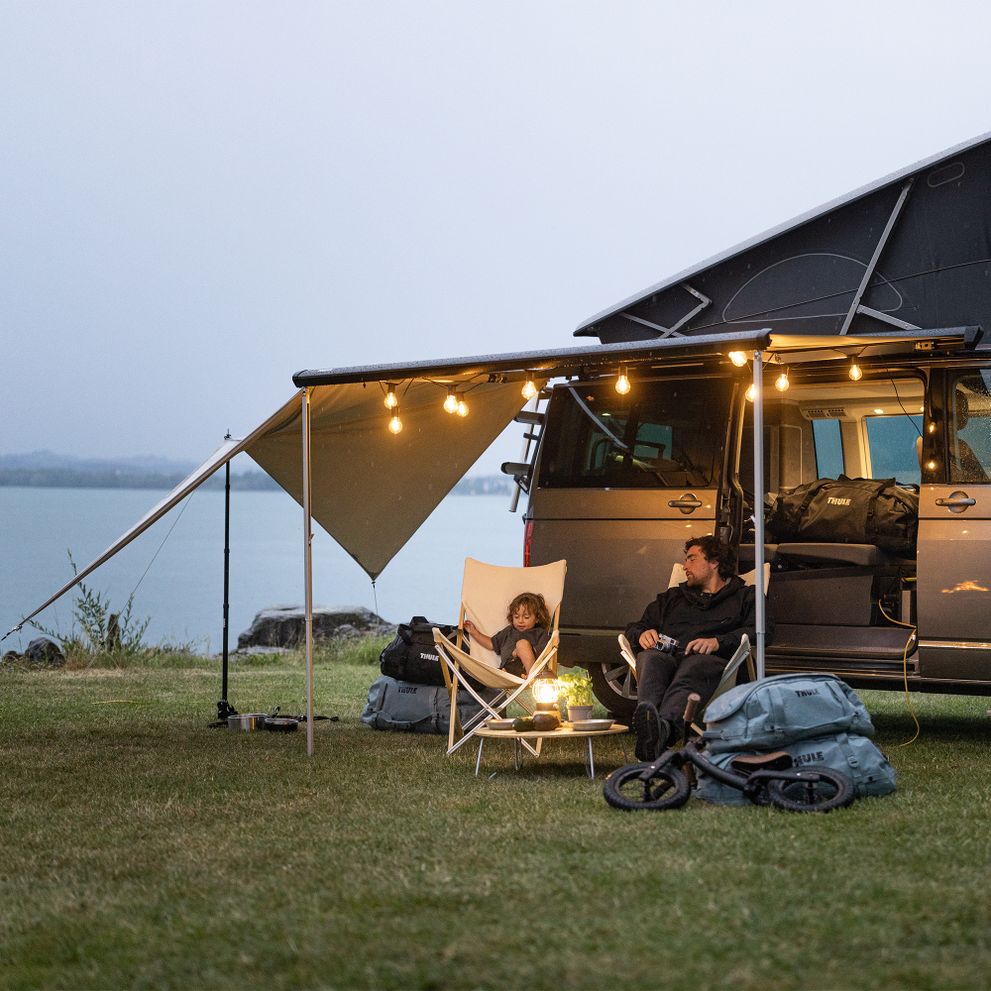 A van is parked by the water with van Thule Subsola awning panels and a camp set up with lamps.
