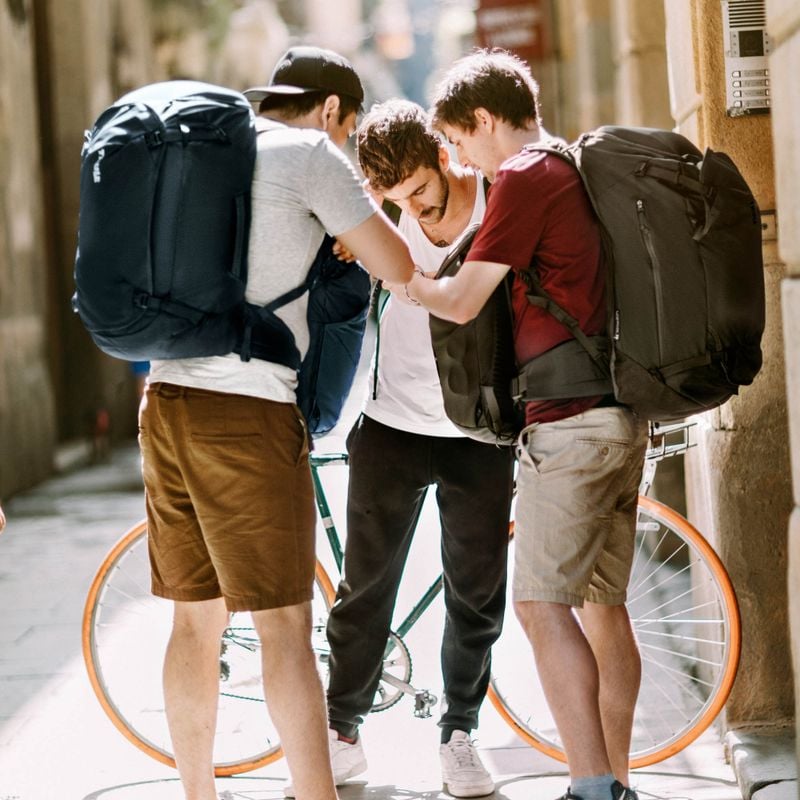 Three backpackers look at a map in a narrow sunny city street wearing Thule Landmark 70L travel backpacks.