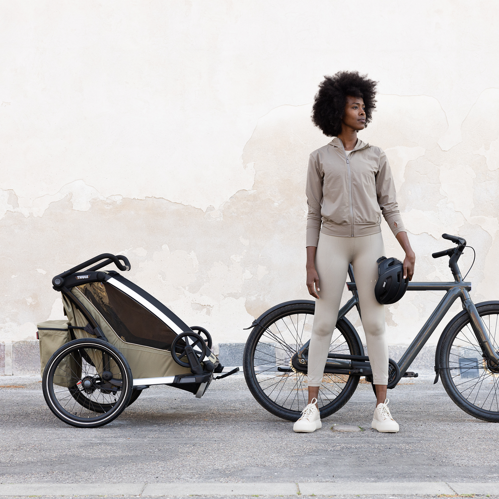 A woman stands by a concrete wall with a bike and a brown Thule Chariot child bike trailer.