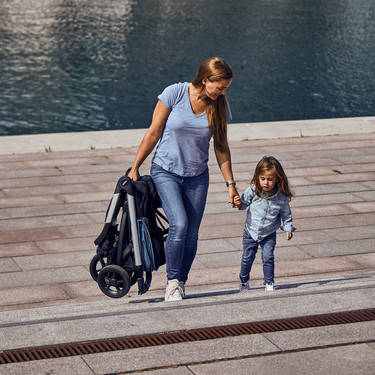 Next to water, a woman walks with her child's hand in one hand, and a folded Thule Spring stroller in the other.