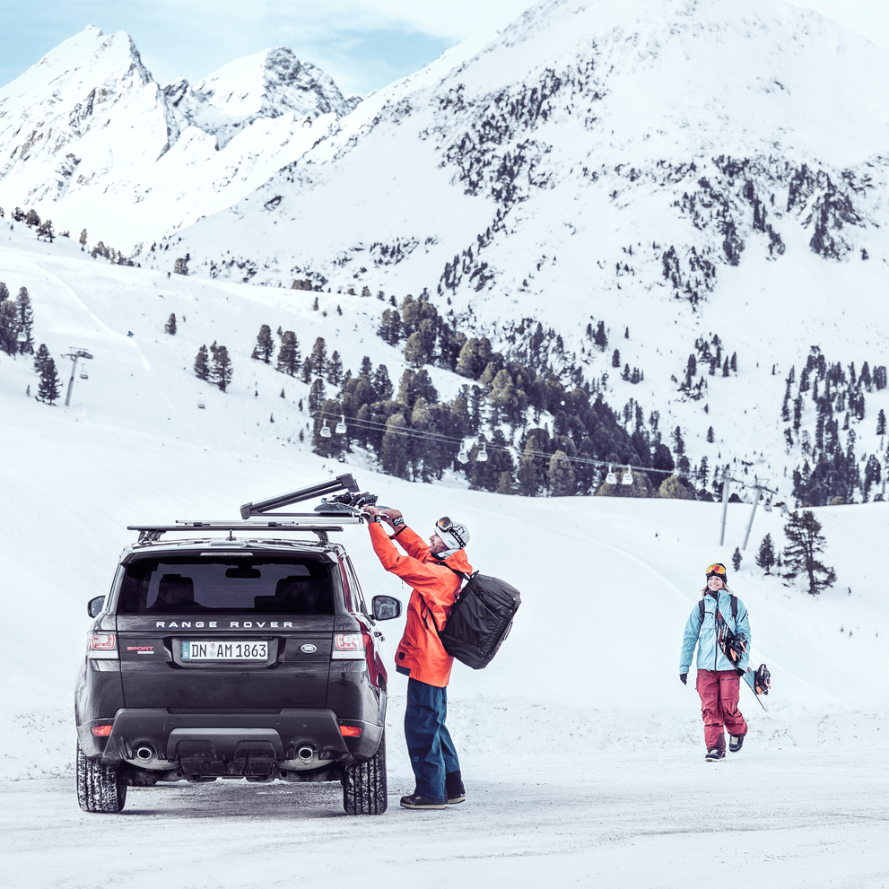 A man is unloading skis from his car roof with a Thule SnowPack Extender while a woman is walking towards him