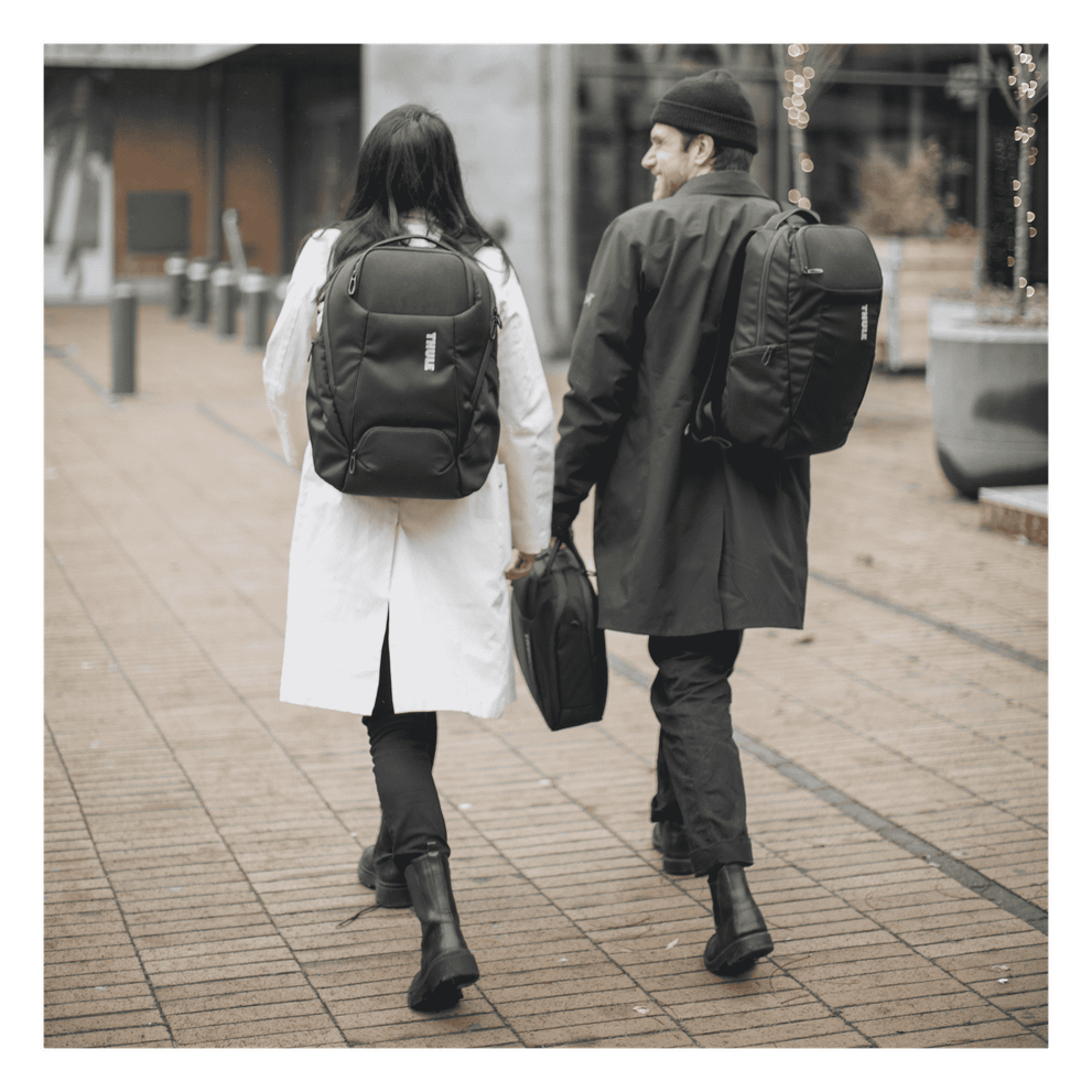 A man and a woman walk towards a glass building, chatting and carrying black Thule Accent backpacks.
