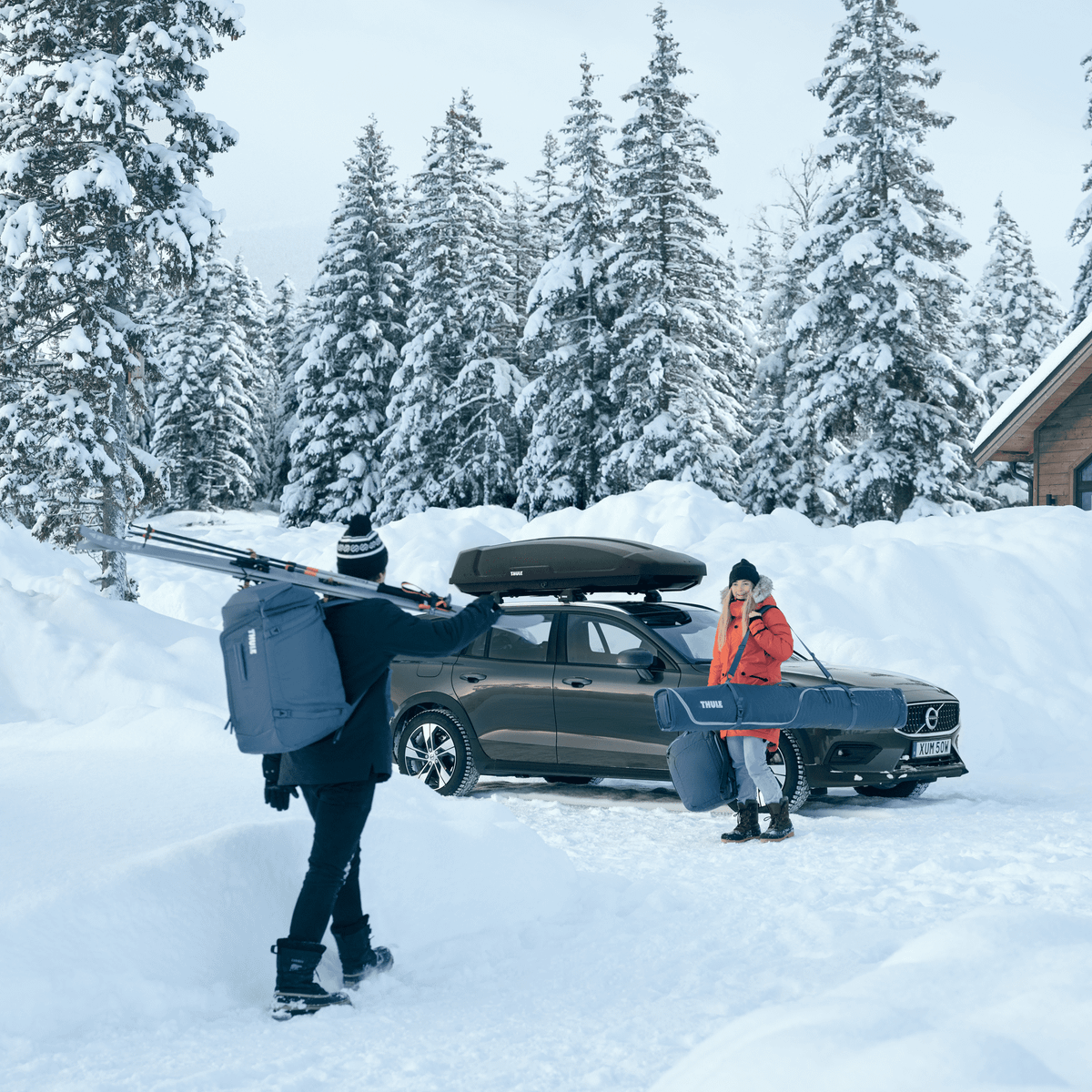 A woman stands by a car in the snow saying hi to a man carrying skis and a Thule RoundTrip 60L Ski Backpack.