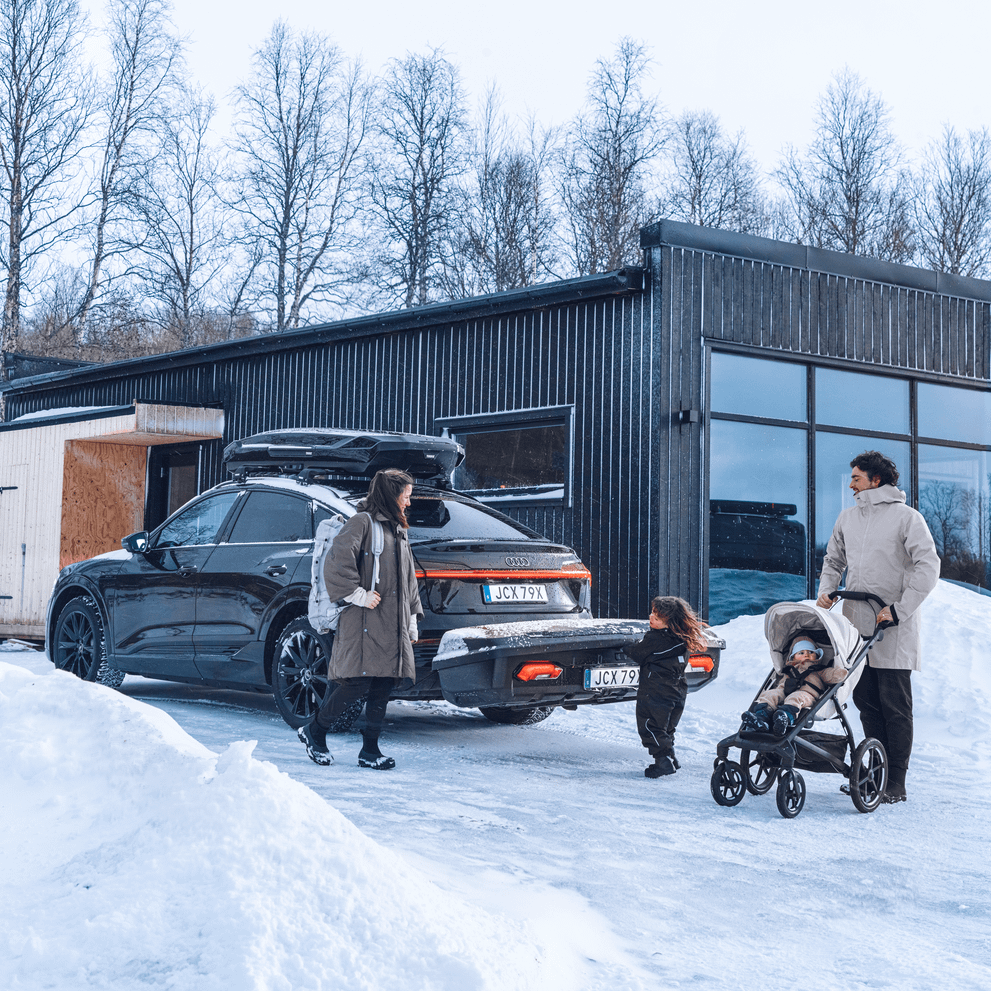 A family with a stroller stand behind their SUV, equipped with both a rooftop box and Thule Arcos XL, outsidee a cabin in a snowy landscape.