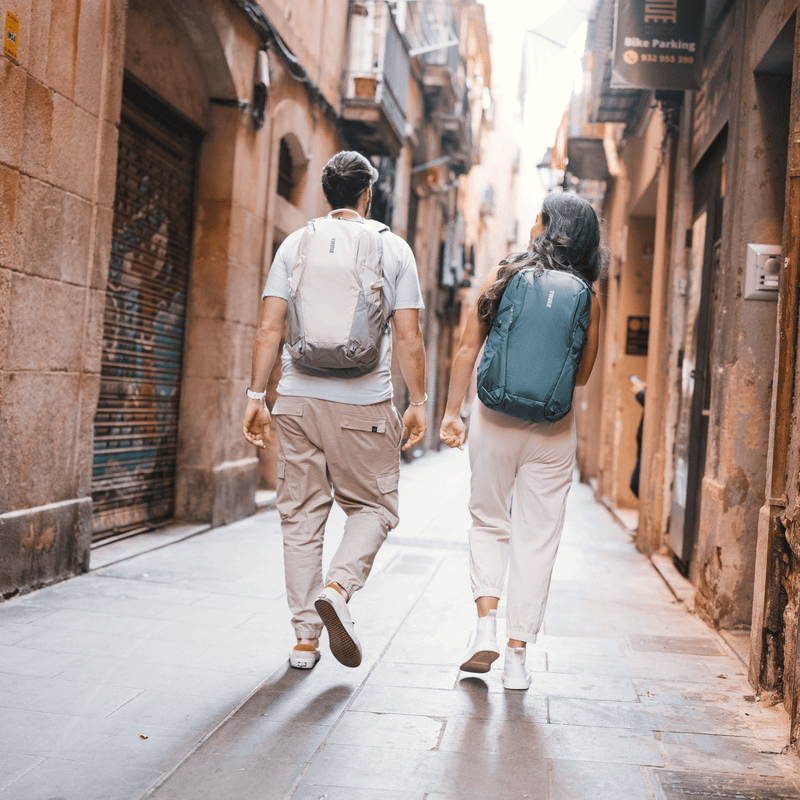 A man and women walk down a sunny, narrow city street with white and green Thule EnRoute backpacks.