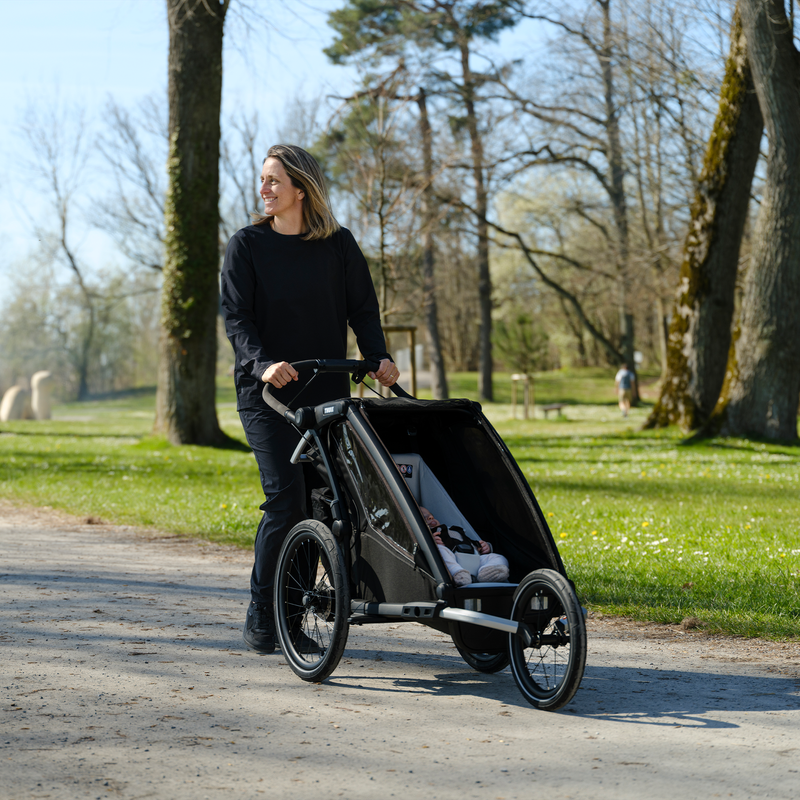 A person pushing a Thule Chariot bike trailer fitted with a Thule Chariot infant sling along a path in a park.
