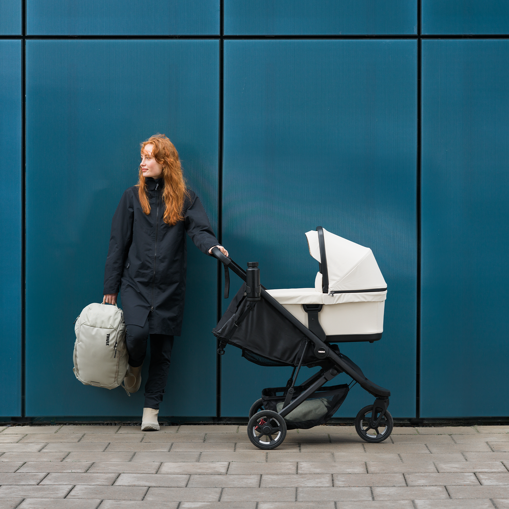 A woman standing in front of a blue wall holding onto the handle of the Thule Spring 2 stroller with a light beige bassinet.