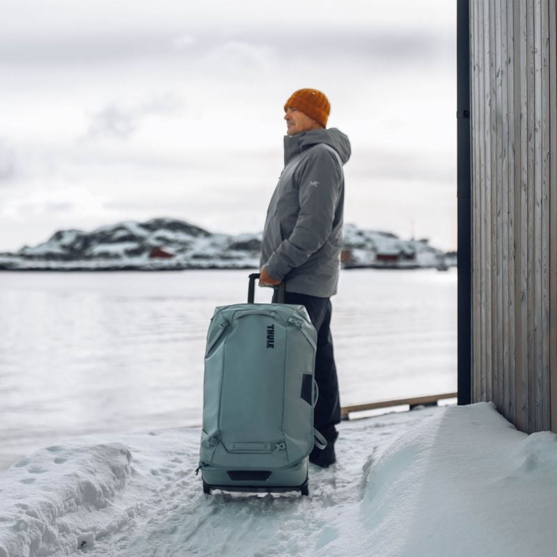 A man stands looking over icy ocean landscape with a blue Thule Chasm check-in suitcase.