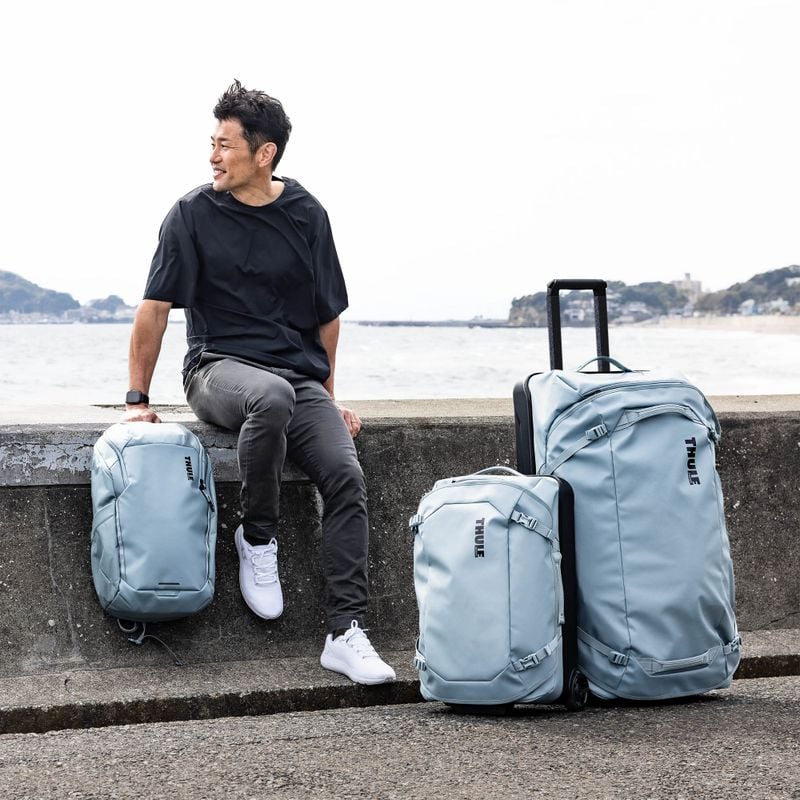 A man sits by the water with blue Thule Chasm backpack, carry-on and check-in suitcase.