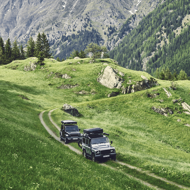 Two cars with roof top tents mounted are driving on a mountain road