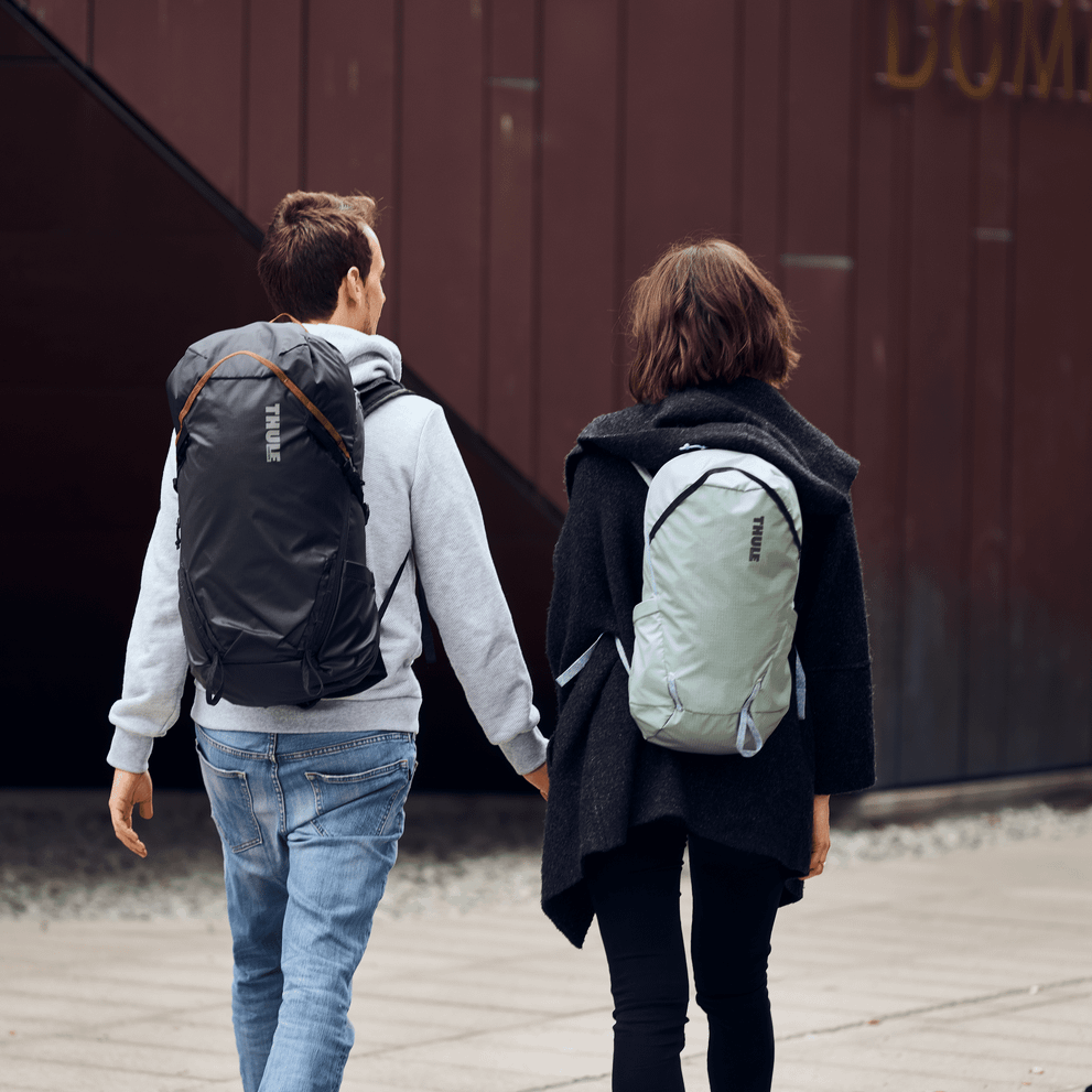 A couple walk next to a rustic building both carrying Thule Stir 35L hiking backpacks.
