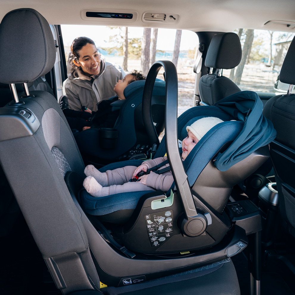 Adult securing a child in a Thule Elm Connected toddler seat inside a vehicle, with another child in a rear-facing car seat beside them.