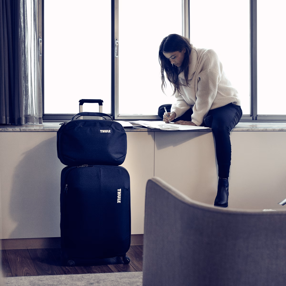 A woman sits on a window sill, with her backpack on a on a Thule Subterra Carry-On Spinner suitcase.