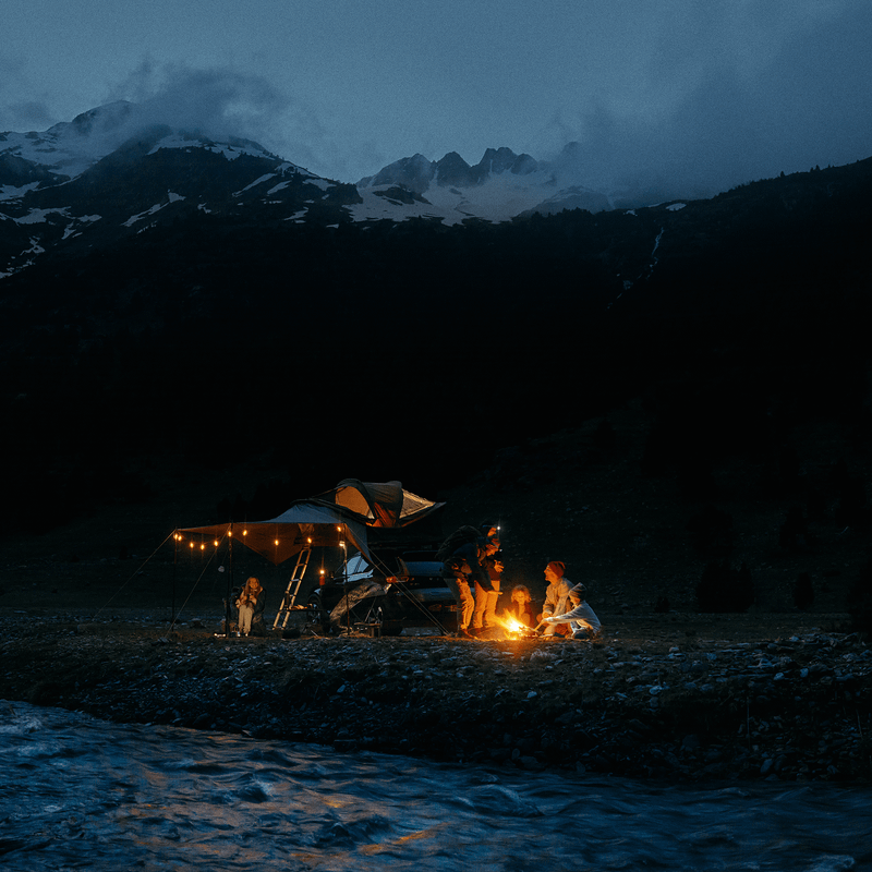 A group of people sitting by a fire beside a car with a roof top tent