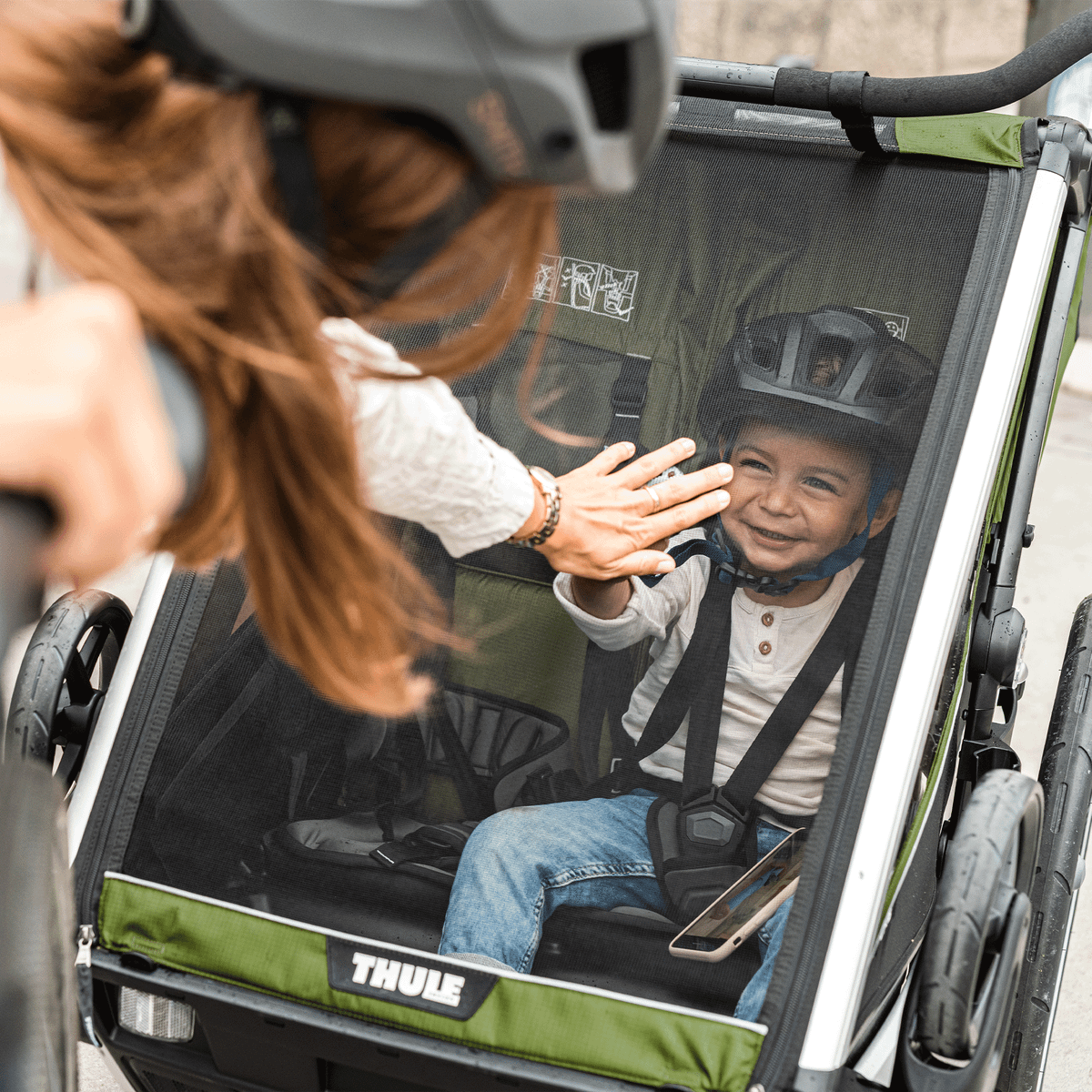 A little boy inside a Thule Chariot Cab child bike trailer high fives his mother.