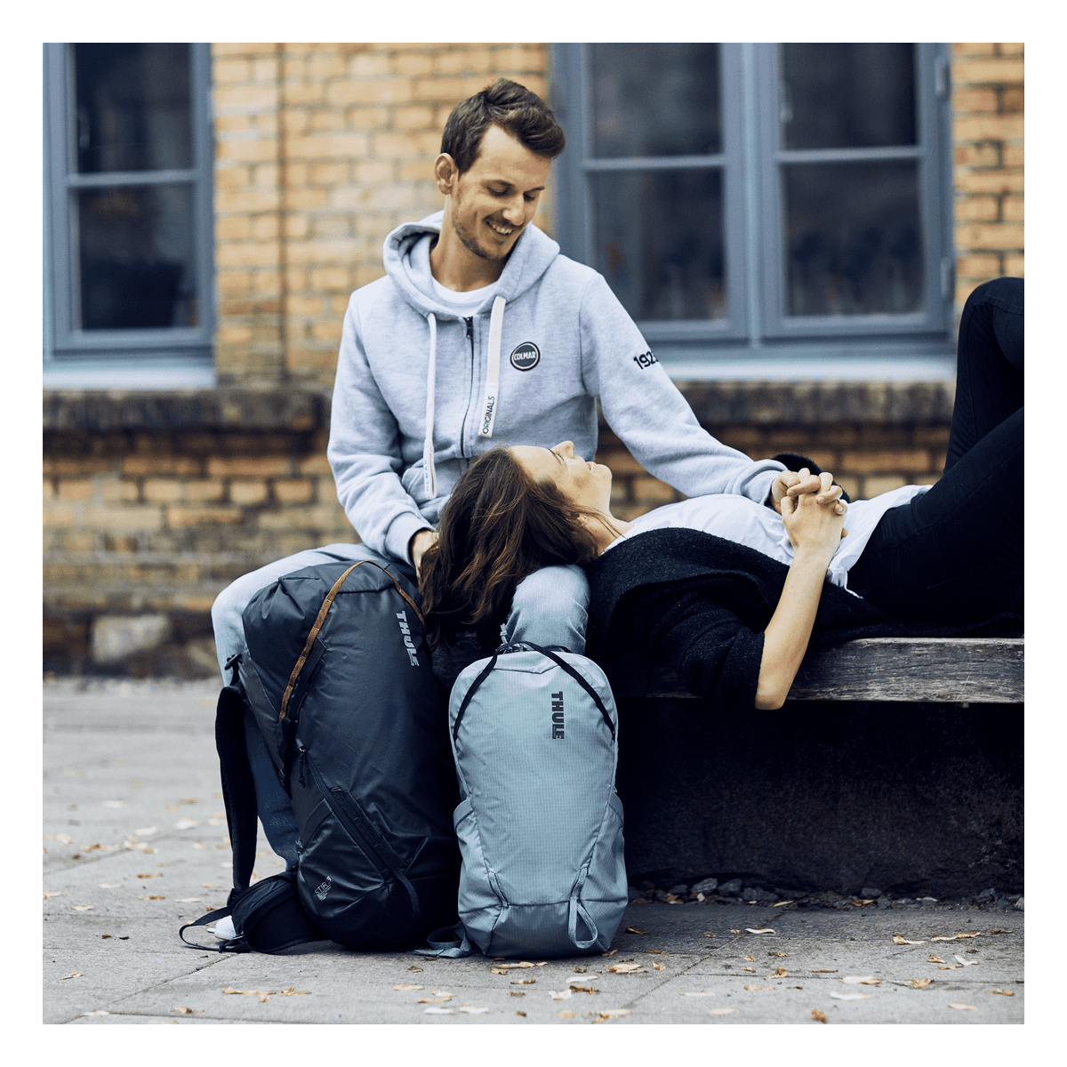On a bench, a woman rests her head on the lap of a man who is holding a black and blue Thule Stir 35L backpack.