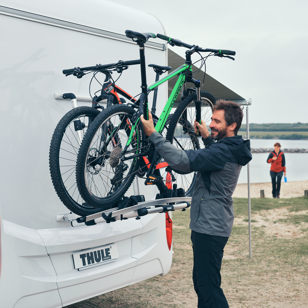 A man unloads bikes from a Thule Excellent Standard van bike rack next to a lake.