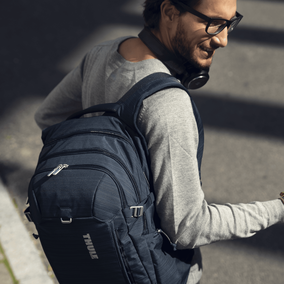 A man carrying headphones crosses a street carrying a blue Thule Construct Backpack.