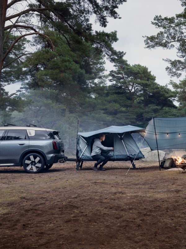An einem Fahrzeug angebrachter Zeltaufbau des Typs Thule Outset auf einem Waldcampingplatz, dargestellt mit Zubehör wie Plane und Campingausrüstung