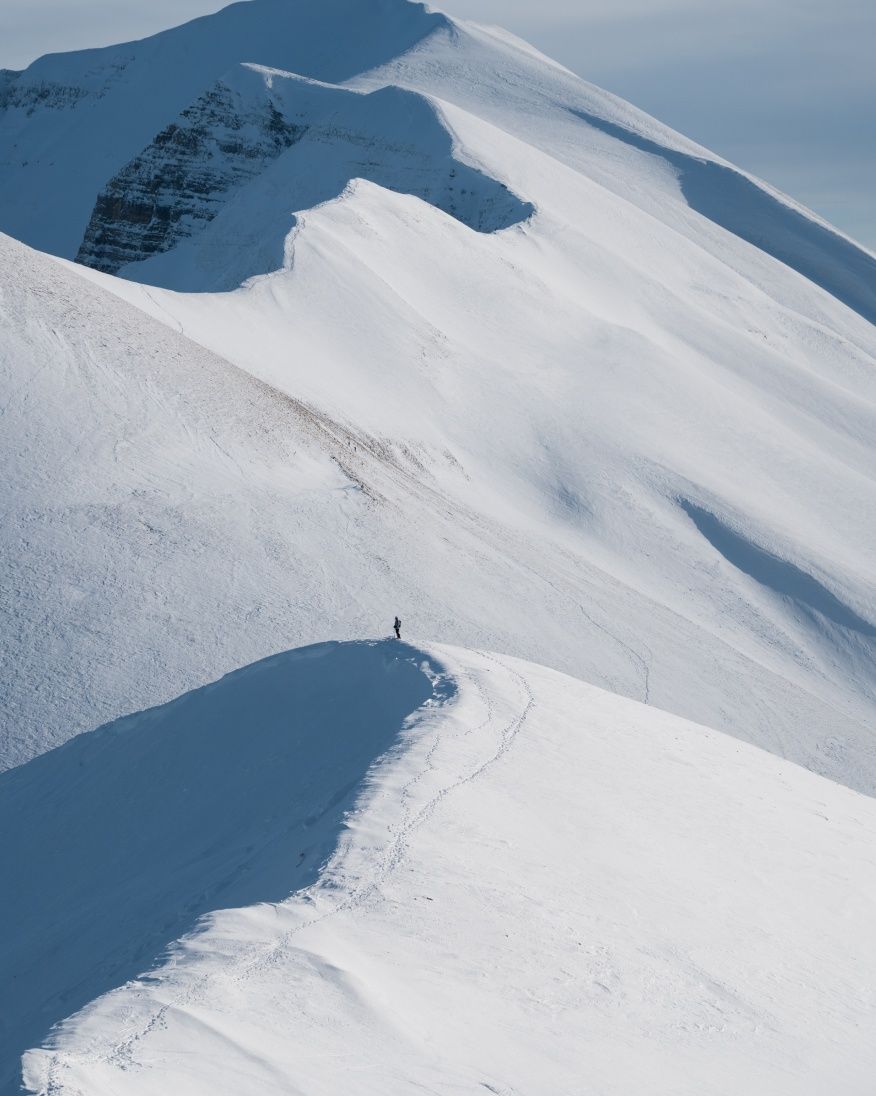 A person far away stands on a snowy cornice while looking out over the mountains.