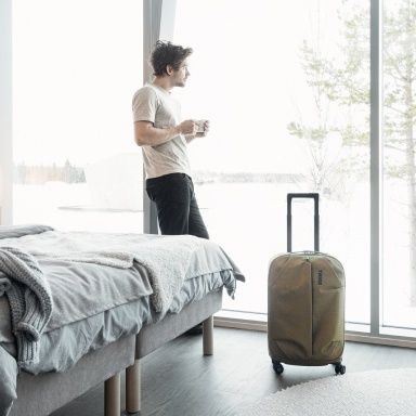 A man in a hotel room looks out the window with a coffee and a suitcase beside him.