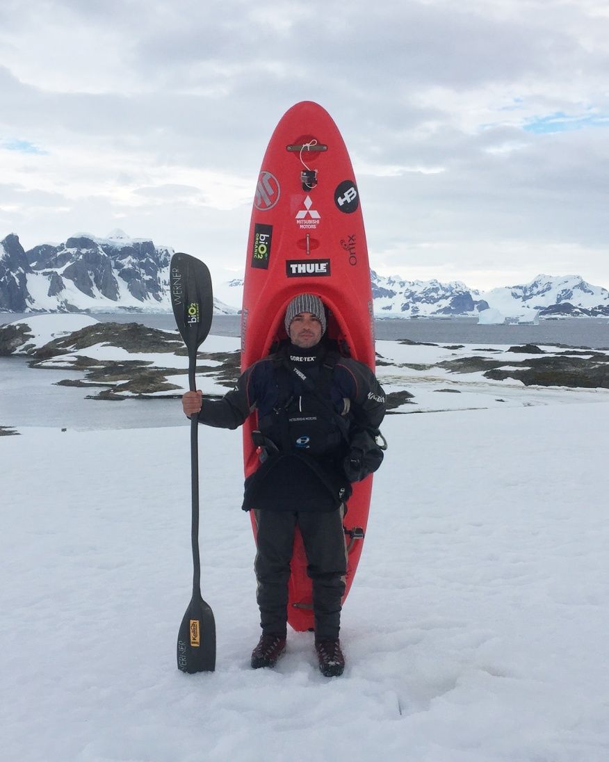 Pedro Oliva stands in the snow  holding a paddle with a red kayak standing behind him.