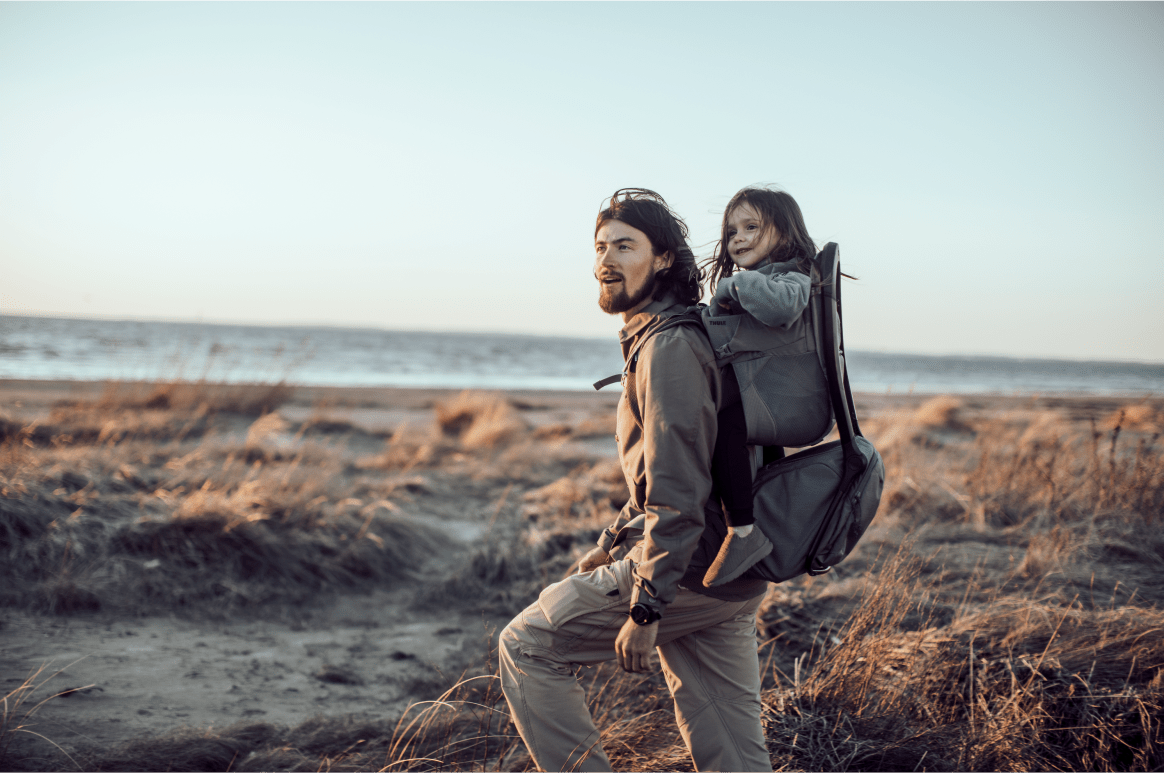 A man walks on the beach with a girl in a Thule child carrier backpack on his back.