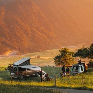 Le persone sono sedute su sedie accanto a due veicoli dotati di una tenda da tetto Thule e una tenda da gancio traino Thule Outset nella natura.
