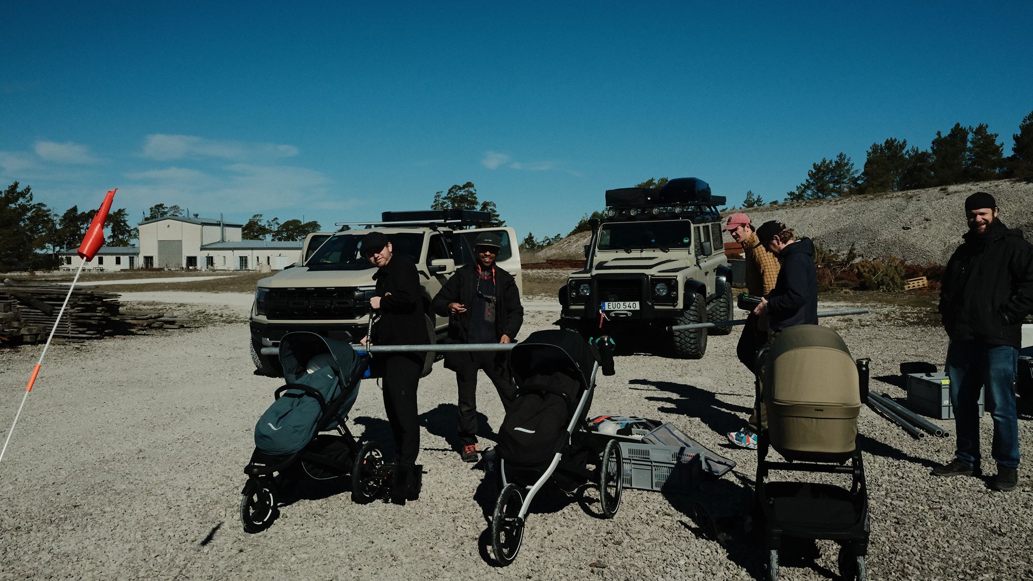 Nos bastidores da campanha A Different Kind of Strong da Thule, com membros da equipe, vários carrinhos de bebê Thule, equipamentos de produção e dois veículos reunidos em uma área de cascalho.