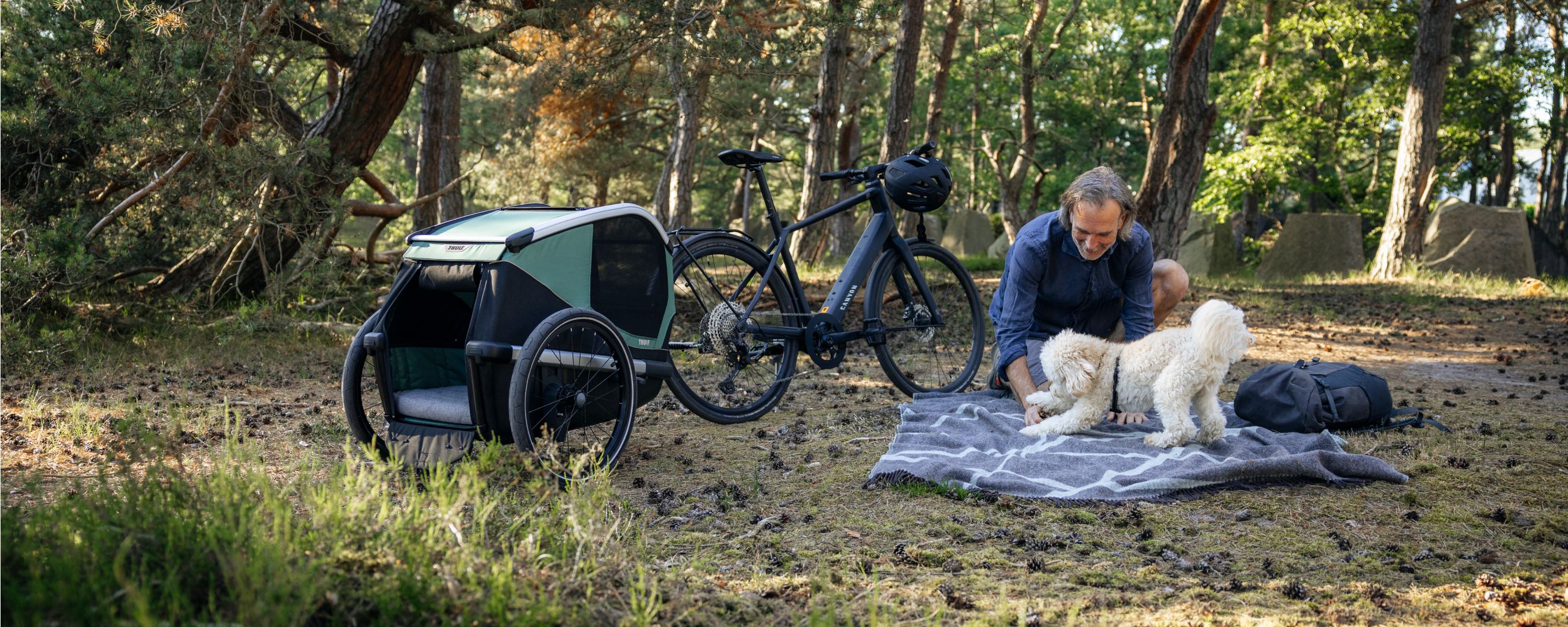 Pessoa a brincar com um pequeno cão branco num ambiente de floresta ao lado de uma bicicleta com um atrelado para cães Thule Bexey acoplado.