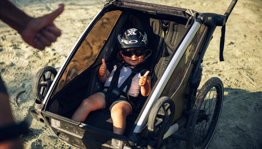 Child seated inside a bike trailer on sand, wearing a helmet and giving thumbs up, with an adult’s hand also visible.