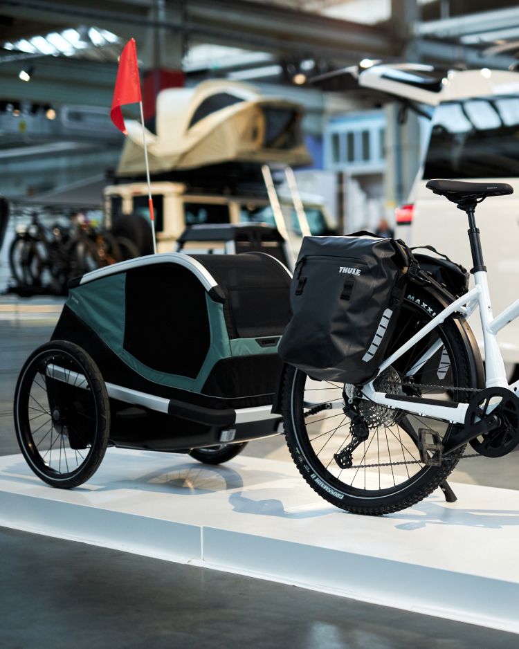 A white bicycle parked on a white platform in an exhibition hall with a Thule Bexey dog bike trailer in dark green and black colors attached to the back of it. The bike trailer has a red flag on it and the bicycle also has two Thule Shield Pannier bags on the back side.