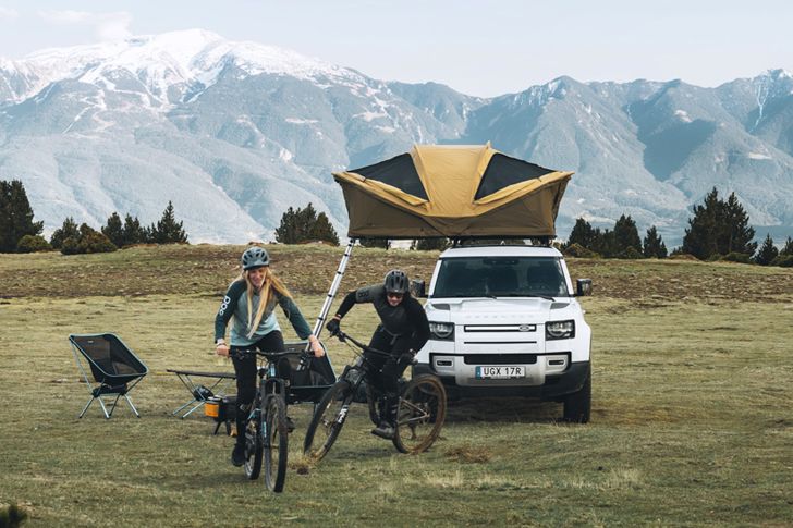 Deux personnes s’éloignent à vélo d’un véhicule stationné sur lequel est montée une tente de toit à coquille souple dans des montagnes enneigées.