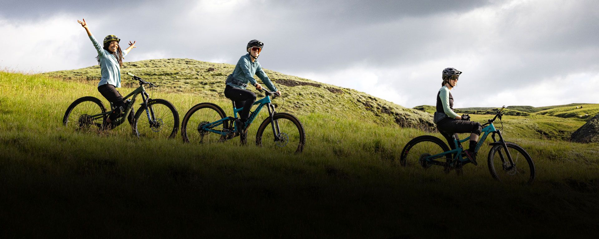 Two cyclists cycle down a grassy hill while one cyclist stands at the top holding out her arms.