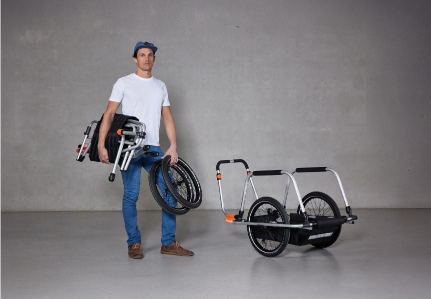 Un homme debout dans un studio tenant une remorque vélo Reacha Sport pliée, présentant son design léger et compact.