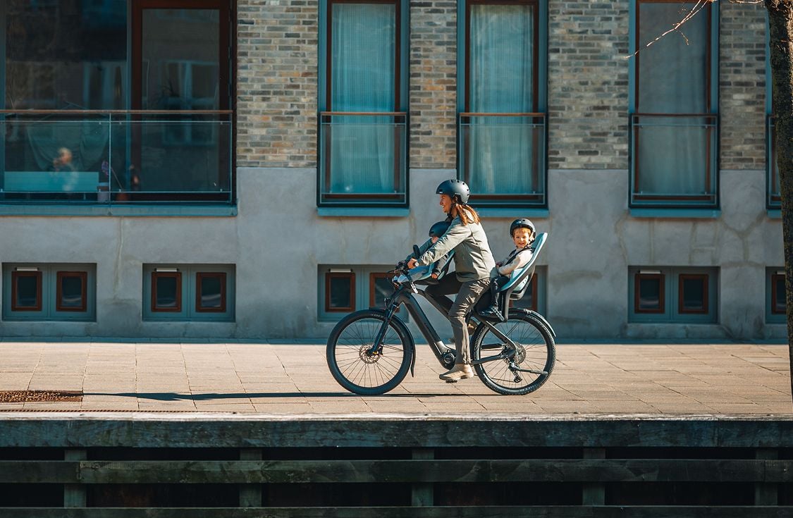 Person riding a bicycle with children in both a Thule Yepp 2 mini and Thule Yepp 2 maxi child bike seats along a waterside path in front of a brick building.